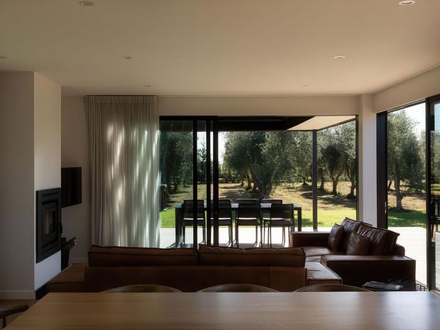 A contemporary open-plan living and dining area with a large timber dining table in the foreground, brown leather sectional seating, a built-in fireplace and television wall, and expansive black-framed glazing opening to an outdoor dining terrace and landscaped olive grove. Neutral finishes and recessed ceiling lighting create a warm, understated interior.