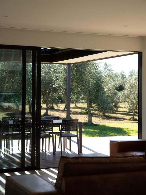 A contemporary interior living area opens onto a covered outdoor deck through extensive dark-framed glazing and sliding doors. A brown leather sofa occupies the foreground, while a dark outdoor dining table and chairs sit on the deck. Beyond is a sunlit landscaped garden with mature trees. The space has a clean, indoor-outdoor connection and strong natural light.