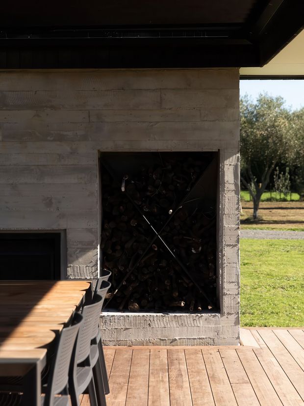 Outdoor entertaining area with a built-in concrete fireplace and recessed firewood storage, opening onto a timber deck and rural landscaped surroundings. A dark timber dining table and modern outdoor chairs occupy the foreground.