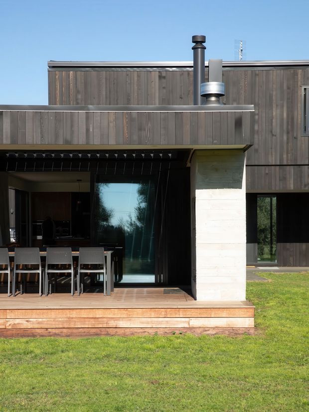 Exterior view of a contemporary two-storey home with a dark timber-clad upper facade, broad metal roof and projecting canopy. The ground level opens onto a timber deck with an outdoor dining table and chairs, large glazed doors, and a pale masonry column supporting the canopy. A chimney and metal flue are visible on the roof, with lawn in the foreground.