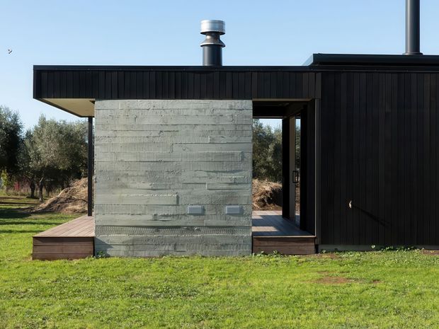 Exterior view of a compact contemporary house in a rural landscaped setting. The building features a flat roof, black vertically ribbed cladding, a rough exposed concrete block or rendered central wall, dark-framed openings, and a small timber deck or entry platform. A chimney and roof flue are visible above the roofline.