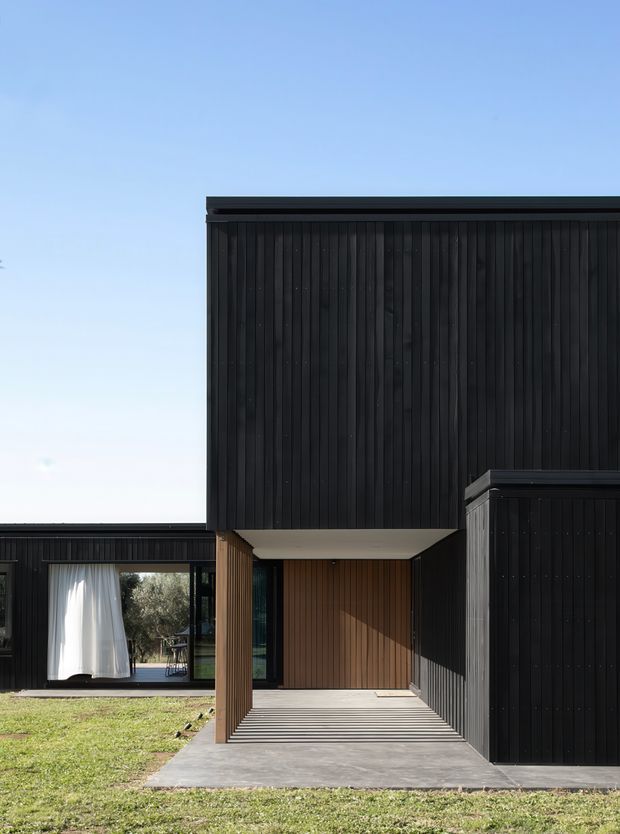 Contemporary residential exterior featuring a bold cantilevered upper volume clad in dark vertical timber boards. A recessed entry porch with warm timber lining and horizontal decking sits beneath the overhang, beside large glazed openings with white curtains. The low, boxy composition has a flat roof and strong contrast between black cladding, natural timber, concrete paving, and lawn.