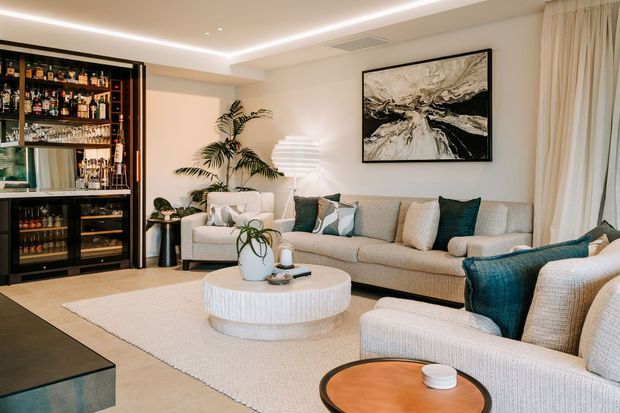 A refined contemporary living room with a large textured sectional sofa, coordinating armchairs, a round white coffee table, soft cream rug, black built-in bar cabinetry, layered sheer curtains, indoor palms, and recessed plus concealed lighting. Teal cushions and upholstery provide contrast against the warm neutral palette, while abstract monochrome artwork forms a feature above the sofa.
