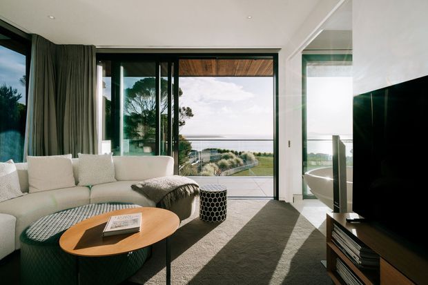 Bright contemporary living room with a large sectional sofa, oval timber coffee table, textured ottoman, and television on built-in joinery. Full-height sliding glass doors open to a balcony or terrace with views toward the water and landscaped coastal surroundings. A freestanding bathtub is visible through the glazed opening on the right, suggesting an adjacent bathroom. The room features a restrained neutral palette, dark window frames, strong natural light, and a timber-lined exterior ceiling.