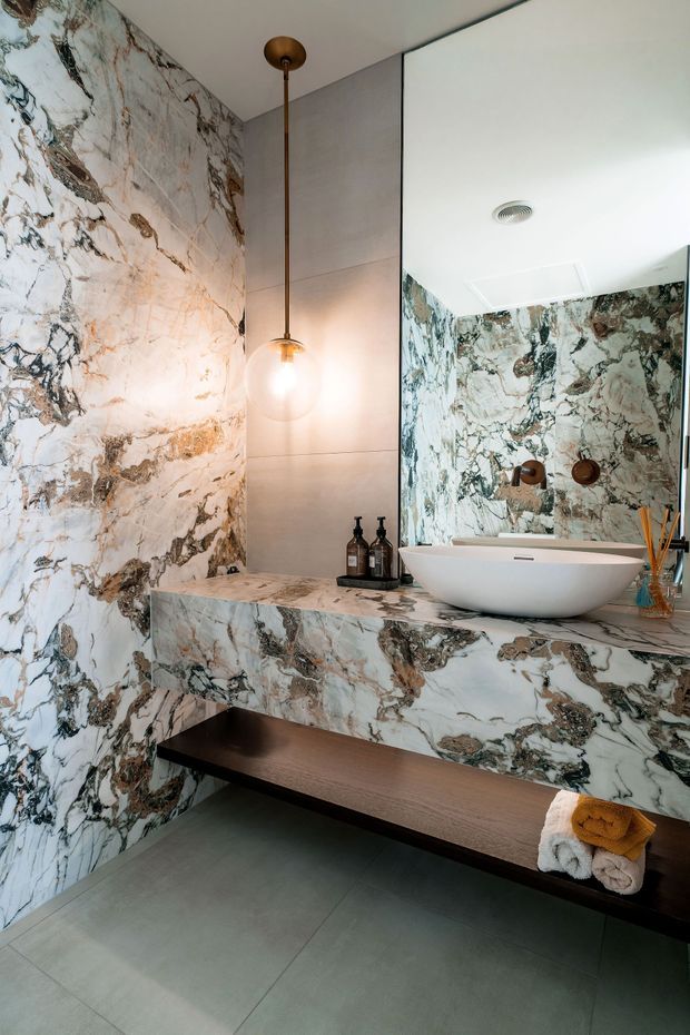 A luxurious contemporary bathroom featuring a dramatic veined marble wall and matching marble-clad vanity. A white vessel basin sits on the floating vanity, with a large mirror, brushed metal wall panel, pendant globe light, dark timber open shelf, and large grey floor tiles.