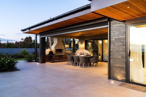 A contemporary covered outdoor entertaining area adjoining a modern home. The expansive patio features pale large-format paving, a timber-lined soffit, dark steel framing, a stone-clad wall, built-in fireplace with wood storage, and a dining table with woven chairs. Glazed doors connect the patio to the interior, while landscaped planting is visible at the edge.