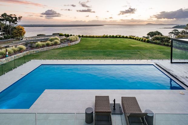 A landscaped waterfront property viewed from an elevated terrace, with a large rectangular swimming pool in the foreground, pale paved surrounds, glass balustrades, and two outdoor loungers. Beyond the pool is a broad manicured lawn bordered by curved stone retaining walls and ornamental planting, opening to expansive coastal water and a distant shoreline under a soft sunset sky.