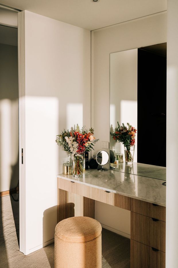 A warm, sunlit dressing or vanity area with a built-in timber vanity desk, pale marble-look benchtop, large wall mirror, drawers, and a rounded upholstered stool. A doorway opens to an adjacent space at left, while flowers and small personal items decorate the vanity.