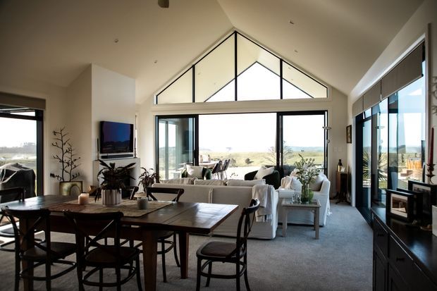 A spacious open-plan living and dining area with a vaulted ceiling and prominent triangular clerestory glazing. The room features large black-framed windows and sliding doors overlooking a rural landscape, a dark timber dining table with cross-back chairs, a light sectional sofa, dark built-in cabinetry, and neutral carpeting. The interior is bright, refined, and connected to the outdoors.