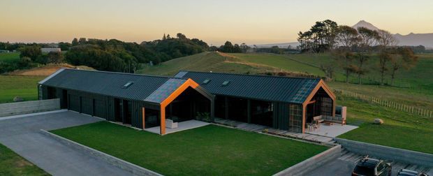 Aerial exterior view of a long, low-slung contemporary rural residence with dark corrugated cladding and gabled roof forms accented by vivid orange trim. The house sits within landscaped lawns and fenced rolling farmland, with distant hills visible beyond.