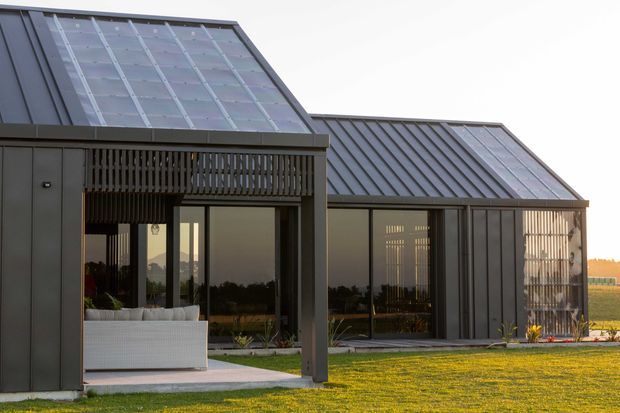 Exterior view of a contemporary single-storey building composed of two connected gabled volumes. The facade uses dark vertical metal cladding, extensive floor-to-ceiling glazing, and timber-like vertical screening beneath the covered outdoor entry or patio. The steep roof planes include broad translucent or glazed sections, and the building opens onto a manicured lawn with agricultural landscape beyond.