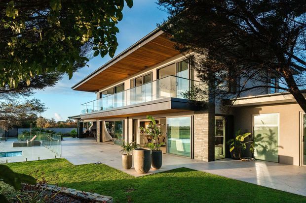 Exterior view of a contemporary two-storey home with broad roof overhangs, a glazed upper-level balcony, extensive floor-to-ceiling glazing, pale masonry walls, and a large paved terrace beside a swimming pool. Landscaped garden and mature trees surround the house.