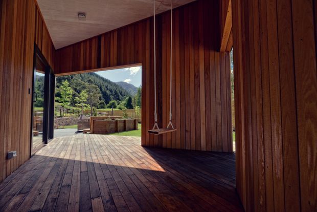 A warm timber-lined indoor space opening directly onto a spacious timber deck and outdoor seating area. Vertical wood wall panelling, dark timber flooring, exposed timber structure, and large openings frame a lush mountainous landscape. Two hanging swing seats add a playful feature to the otherwise minimalist retreat-like setting.