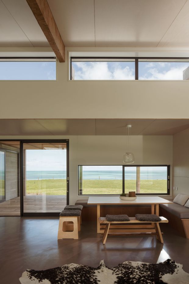 A bright coastal living and dining area with expansive black-framed glazing overlooking a grassy shoreline and ocean. The room features a built-in window-side bench with cushions, a timber dining table and bench seating, a compact upholstered stool, polished dark flooring, warm neutral walls, a suspended glass pendant light, and a prominent exposed timber ceiling beam. High clerestory windows and a partially double-height ceiling add natural light and openness.