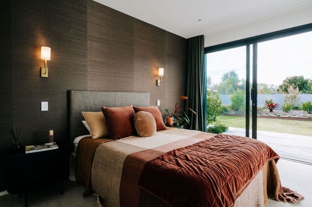 A refined contemporary bedroom with a large upholstered bed dressed in layered brown, rust, taupe and cream textiles. The room features a dark textured feature wall, matching dark bedside furniture, warm wall sconces, full-height dark curtains and expansive sliding glass doors opening to a landscaped outdoor area and timber deck.