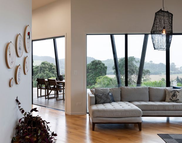 Bright contemporary living room with a large L-shaped upholstered sectional sofa, timber flooring, expansive black-framed glazing, and a woven pendant light. A dining area is visible through the opening to the left, while decorative circular wall pieces and indoor plants add detail. The glazing overlooks a green, hilly landscape.