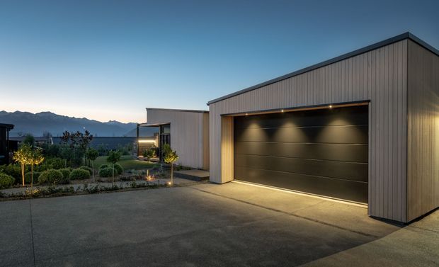 Twilight exterior view of a contemporary detached garage and driveway beside a modern single-storey home. The garage has vertical dark cladding, a broad dark sectional door, concealed linear lighting, and a clean low-pitched roofline. A landscaped garden and illuminated path sit alongside the building, with distant mountains visible beyond the property.