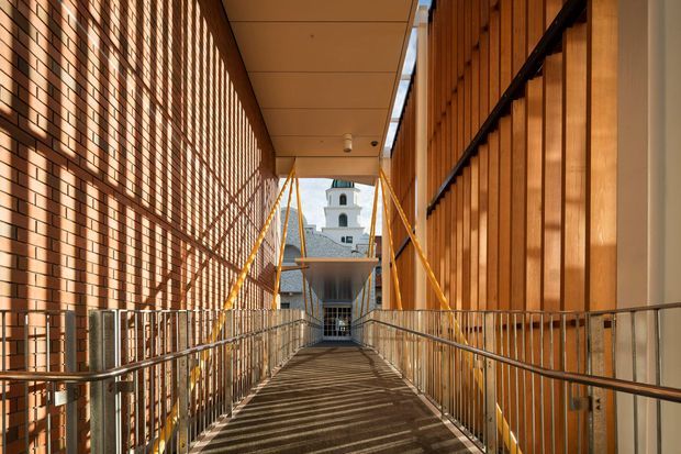 A covered architectural walkway or bridge enclosed between a warm red-brick wall on the left and vertical timber-clad facade on the right. Stainless-steel handrails line both sides, while yellow structural braces and strong sunlight create rhythmic diagonal shadows across the dark walkway. A white tower-like building is visible at the far end.