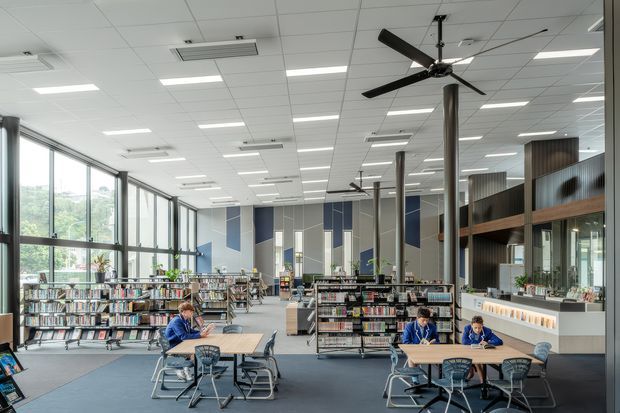 A bright, spacious modern library or educational reading space with extensive bookshelves, study tables and chairs, large floor-to-ceiling windows, exposed dark ceiling fans, acoustic ceiling tiles, and a café or service counter along one side. The interior uses cool neutral tones with blue and timber accents.