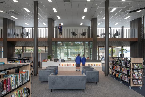 A bright, double-height contemporary library or commercial reading space with rows of bookshelves, grey upholstered lounge seating, a central service or display counter, tall dark structural columns, and an elevated mezzanine walkway with additional seating. Extensive glazing and linear ceiling lighting create an open, institutional interior.