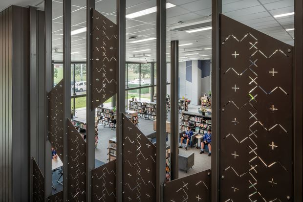 Interior architectural view through tall dark metal screens with perforated geometric patterns, overlooking a bright multi-level library or commercial reading space. Floor-to-ceiling glazing, exposed structural columns, bookshelves, seating, and a suspended grid ceiling are visible. The image emphasizes the decorative screening and open institutional interior.