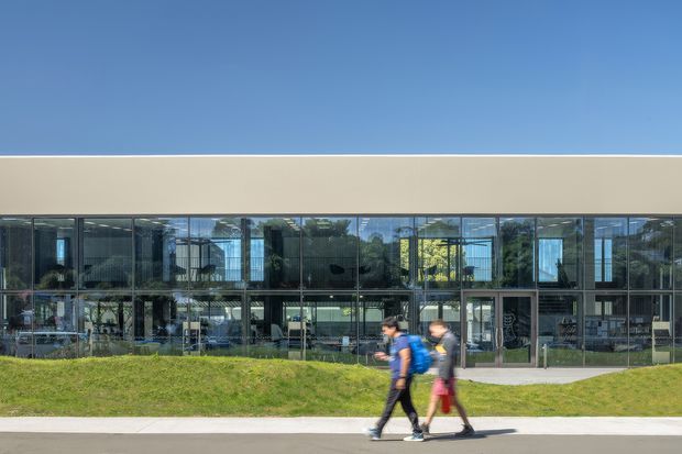 Wide exterior photograph of a contemporary two-storey glazed building with a long, low rectangular form. The upper level has a plain light beige facade band, while the lower and upper elevations feature extensive floor-to-ceiling glass and dark metal framing. A landscaped grassy verge, paved walkway, and pedestrians sit in the foreground under a clear blue sky.