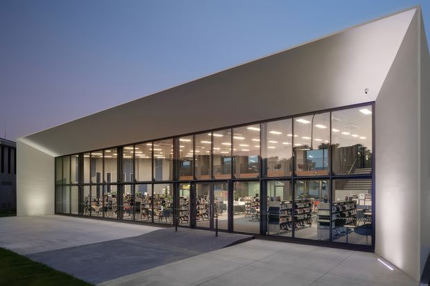 Exterior view of a contemporary commercial building with a dramatic angular, sloping roof canopy and extensive floor-to-ceiling glazed curtain walls. The illuminated interior appears to contain retail shelving, tables, and seating, while the foreground is a paved entry area. The facade is composed of smooth light-colored panels and dark metal-framed glazing, photographed at dusk.