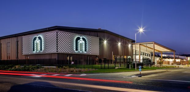 Twilight exterior photograph of a contemporary commercial building on a corner site. The long dark-clad facade features a prominent chevron-patterned panel with two illuminated arched windows, while a glazed entrance canopy projects at the right. Street lighting, fencing, paving, landscaped planting, and vehicle light trails emphasize the urban streetscape setting.