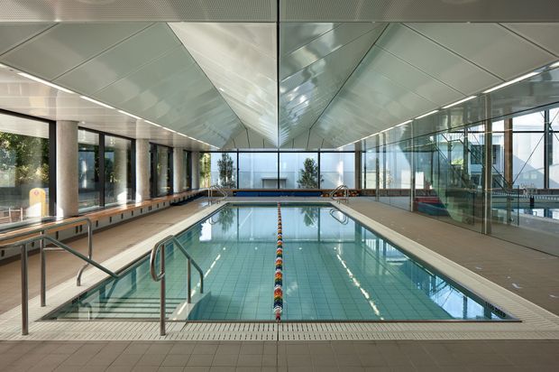 A large indoor swimming pool in a contemporary glazed aquatic facility. The rectangular lap pool has marked swimming lanes, stainless-steel handrails, tiled surrounds, timber-toned bench seating, extensive floor-to-ceiling glazing, and a reflective faceted ceiling that creates a bright, spacious atmosphere.