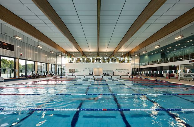 Large indoor aquatic centre with a competition-style swimming pool divided into multiple marked lanes. The bright, expansive hall has a gridded suspended ceiling, prominent exposed timber beams, extensive glazed walls, tiled pool surrounds, spectator seating, and a mezzanine level with viewing areas.