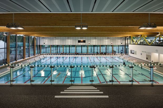 Wide interior view of a large modern indoor aquatic centre, looking across multiple swimming lanes from a raised spectator or circulation area. The pool hall has expansive glazing, timber-clad structural beams, a patterned suspended ceiling, bright overhead lighting, tiled pool surrounds, lane ropes, starting blocks and spectator seating.