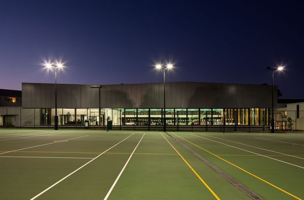 Night-time exterior view of a contemporary institutional or commercial building behind a large illuminated multi-court sports surface. The building has a long, low rectangular form with dark vertical cladding, extensive ground-level glazing, and bright interior lighting. Tall floodlights illuminate the marked green courts in the foreground.
