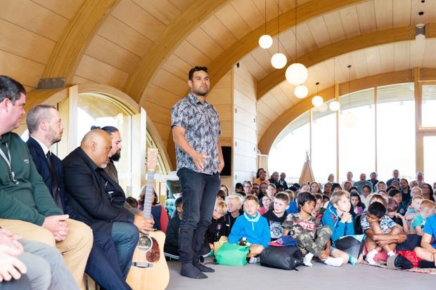A bright communal interior with a dramatic curved timber-lined ceiling, exposed arched timber beams, and large arched windows. A speaker stands at the front addressing a seated audience, with several people and guitars visible in the foreground. The space appears to be a large multipurpose hall or community gathering room.