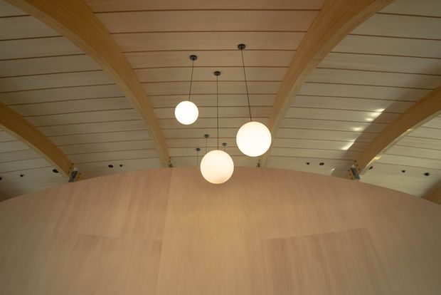 Interior architectural view looking upward at a warm timber-lined ceiling with large curved exposed timber beams and a sweeping curved timber wall or partition. Three suspended spherical pendant lights form the central focal point, with additional small recessed or surface-mounted ceiling lights. The composition highlights expressive wood structure and a softly illuminated, sculptural interior.