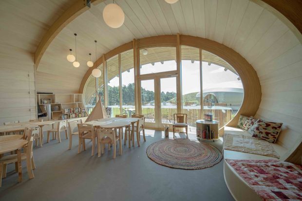 A bright, curved-roof open-plan room with extensive arched glazing overlooking a landscaped outdoor setting. The space combines a dining area with a built-in lounge bench, light timber tables and chairs, shelving, pendant lights, and a circular woven rug. White painted timber wall and ceiling panels contrast with warm timber framing and playful, family-friendly furnishings.