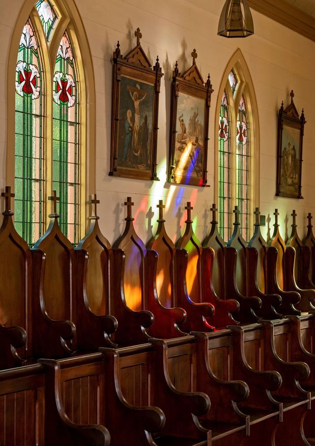Interior view of a historic Gothic-style chapel or church, focused on rows of dark polished timber choir pews with ornate carved finials and armrests. Tall pointed-arch stained-glass windows cast vivid multicoloured light across the timber seating and pale paneled walls. Religious paintings and decorative wall panels line the sides, with a hanging lantern overhead.
