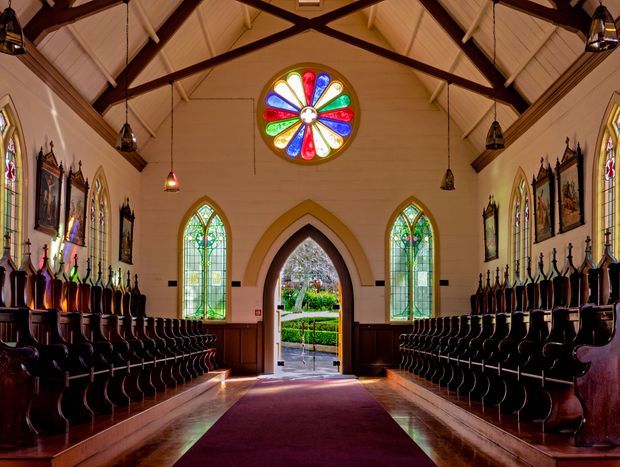Interior view down the central aisle of a historic chapel or church, with rows of dark timber seating, rich timber wall panelling, pointed-arch stained-glass windows, a large circular stained-glass feature window, exposed dark timber roof trusses, pendant lanterns, religious artwork, and a burgundy aisle runner leading to an open arched doorway.