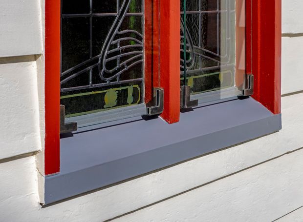 Close-up exterior detail of a rendered or painted white wall with horizontal weatherboard-like lines, a grey window sill, and vivid red-painted timber window frames. The window includes decorative black metal security bars and reflects greenery from outside. The image focuses on facade materials and window detailing.