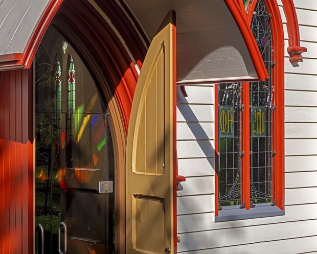 Close-up exterior view of an ornate heritage-style entry, featuring a pointed arched doorway with dark stained glass, a warm timber panelled door, vivid red-painted trims, white horizontal weatherboard cladding, and a narrow leadlight window with decorative coloured glass. The image is taken from outside and focuses on the facade and entrance.