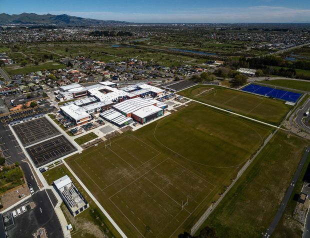 Aerial view of a large modern educational or community campus surrounded by extensive sports fields, including a marked football or rugby field, courts, and running tracks. The low-rise complex has predominantly white roofs with several darker and reddish facade elements, while suburban housing, roads, waterways, and mountain ranges extend across the background.