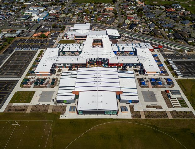 Aerial view of a large contemporary institutional or commercial complex with extensive white flat-roofed building wings, central courtyards, loading and service areas, surrounding car parks, and a sports field in the foreground. Dense suburban development is visible beyond the site.