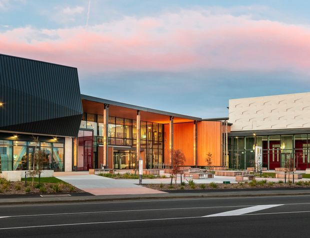 Street-level exterior view of a contemporary commercial building or civic complex at dusk. The facade combines dark ribbed metal cladding, extensive glazed shopfront-style walls, warm timber-toned vertical panels, and a white textured geometric feature wall. A landscaped pedestrian forecourt with young planting, benches, paving, and lighting fronts the building, with a roadway in the foreground.