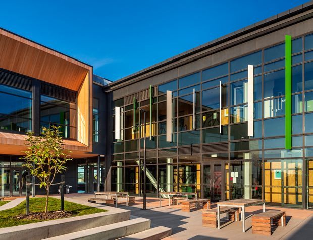 Exterior view of a contemporary multi-storey commercial or institutional building arranged around a landscaped courtyard. The facade combines extensive blue-tinted glazing, dark metal framing and cladding, projecting timber-clad volumes, vertical louvres, and bright green accent fins. The paved courtyard includes concrete seating steps, timber benches and planters, with a glazed entrance and internal stair visible.