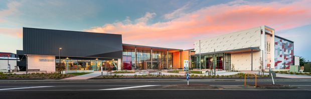 Wide-angle exterior view of a contemporary commercial building complex at dusk, featuring a dark corrugated-metal volume, extensive glazed frontage, a bright orange entry or circulation element, and a white articulated facade with colorful geometric wall accents. The building fronts a paved urban street with landscaped planting and pedestrian access.