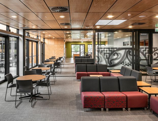 A contemporary commercial lounge and collaborative breakout area with rows of café tables and chairs, modular black and red upholstered seating, glazed partitions, timber wall and ceiling panels, and carpeted flooring. The open-plan interior is brightly lit and includes a bold monochrome graphic on a glass partition.