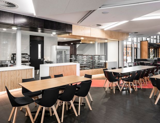 A bright contemporary commercial dining and hospitality interior, likely a café or staff dining space. The room features multiple large timber dining tables with black chairs, a long service counter with vertical timber panelling, a glass-fronted food display counter, commercial kitchen equipment, and integrated cabinetry. White tiled walls, black overhead joinery, dark flooring, exposed services, linear ceiling lighting, and extensive internal glazing create a clean, modern fit-out.