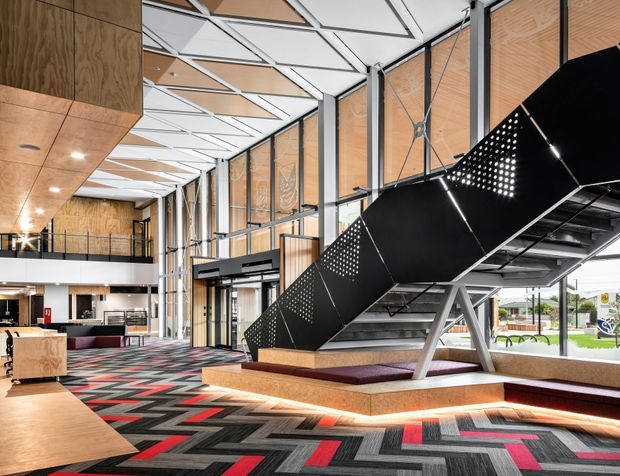 A large contemporary commercial interior with a dramatic black sculptural staircase, exposed steel supports, expansive glazed walls, and a high geometric ceiling. Warm timber wall and ceiling panels contrast with white ceiling elements, while patterned charcoal, grey, and red carpet tiles define the open floor. A reception or lounge area is visible in the background, with daylight entering through the glazing.