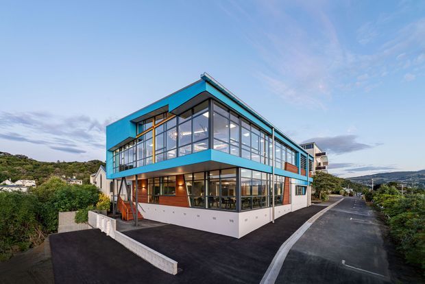 Exterior view of a contemporary multi-storey building on a sloping suburban site, with extensive floor-to-ceiling glazing, bright turquoise-blue cladding bands, dark structural framing, and a paved driveway wrapping around the building. Hillside residential surroundings are visible beyond the property.