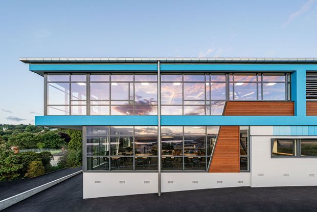 Exterior view of a contemporary multi-level building with a long flat roof, vivid blue horizontal framing, extensive floor-to-ceiling glazing, and warm timber-clad geometric wall sections. The lower level appears to contain an open commercial or communal interior visible through the windows, with a landscaped suburban setting beyond.