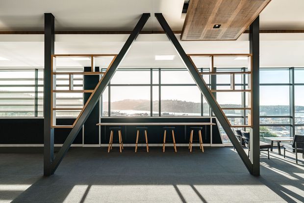 A bright contemporary commercial interior with a dramatic black steel A-frame structural feature, open timber-framed shelving or partitions, and a long built-in black counter with timber stools. Floor-to-ceiling glazing provides expansive views over a waterfront or coastal urban landscape. The space has dark carpet flooring, exposed structural elements, and a distinctive timber slatted ceiling panel.